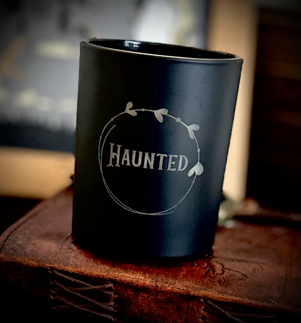 Haunted gothic candle - matt black jar with etched occult designs, scented with dark honey & tobacco, emitting eerie glow.