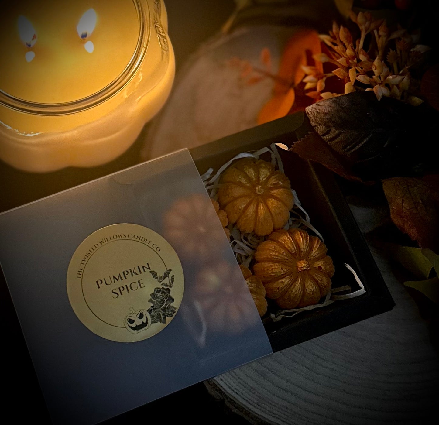 A boxed set of 4 mini pumpkin wax melts scented with dark pumpkin spice amidst autumn foliage