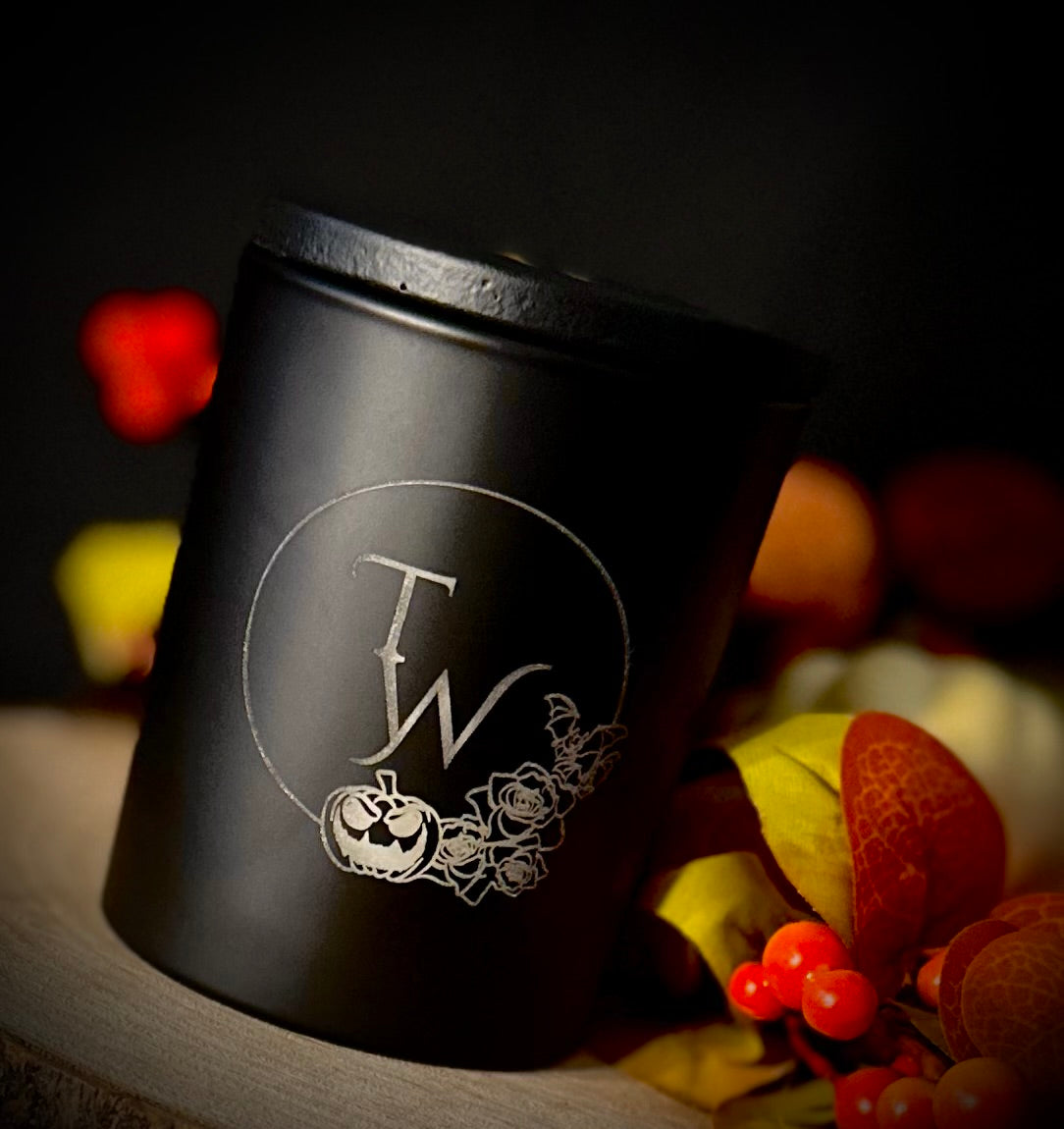Black etched candle jar with bat, pumpkin, and rose design next to dried autumn leaves.