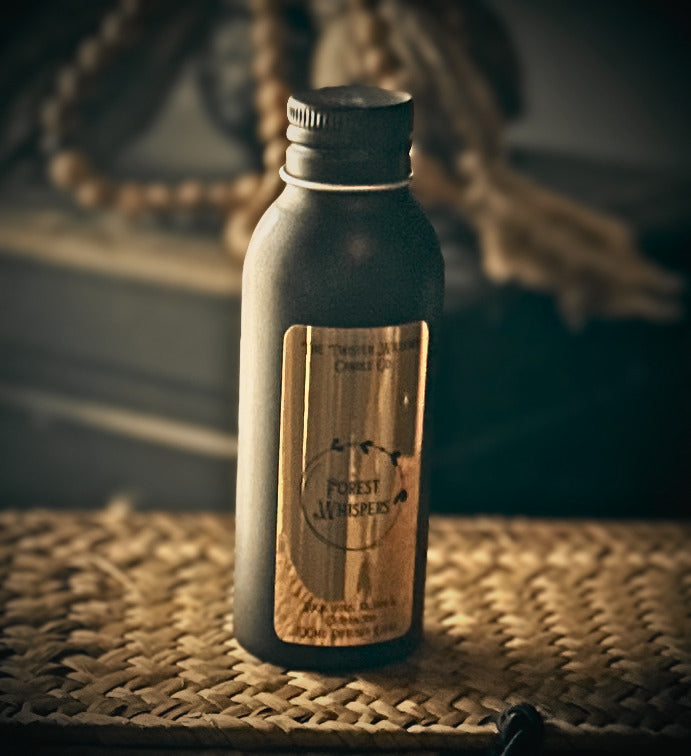 A black metal bottle with a black cap and a gold label reading 'Forest Whispers', placed on a woven mat with a rustic, blurred background.