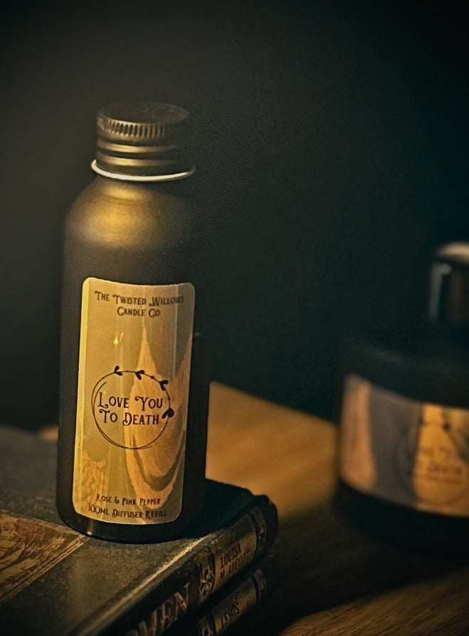 A black metal bottle with a gold label reading 'Love You To Death' by The Twisted Willow Candle Co, styled on vintage books against a dark background.