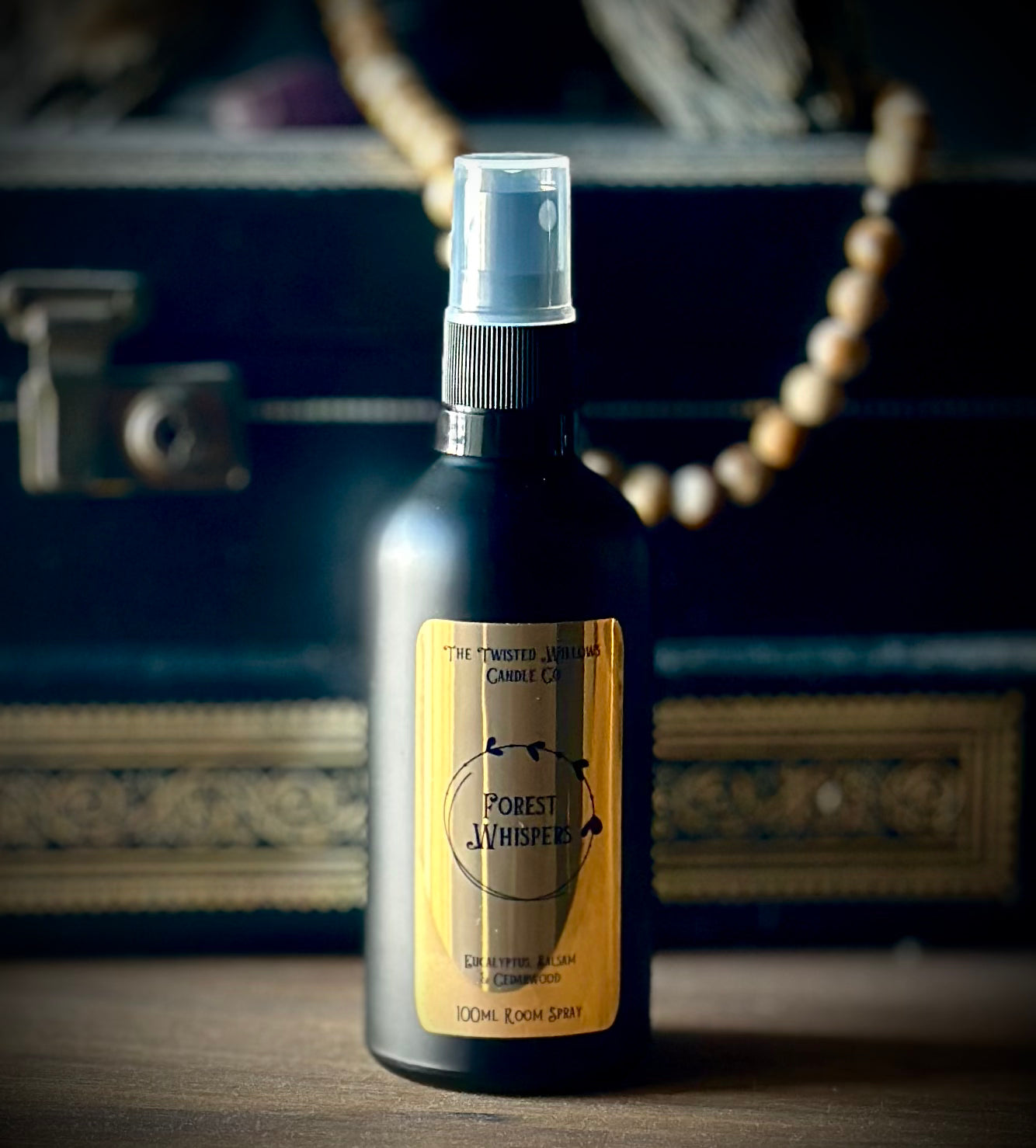 A 100ml spray bottle of Forest Whisper Deodorant by The Twisted Jar Candle Co, featuring a black bottle and a gold label, set against a dark vintage suitcase background with a wooden bead necklace.