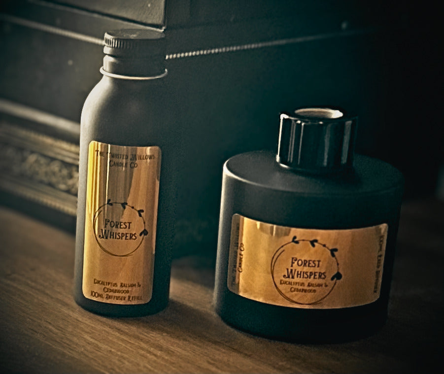 Two Forest Whispers diffuser products on a wooden surface in a dimly lit setting: a tall matte black refill bottle with a gold label on the left and a larger matte black reed diffuser bottle with a black cap on the right.