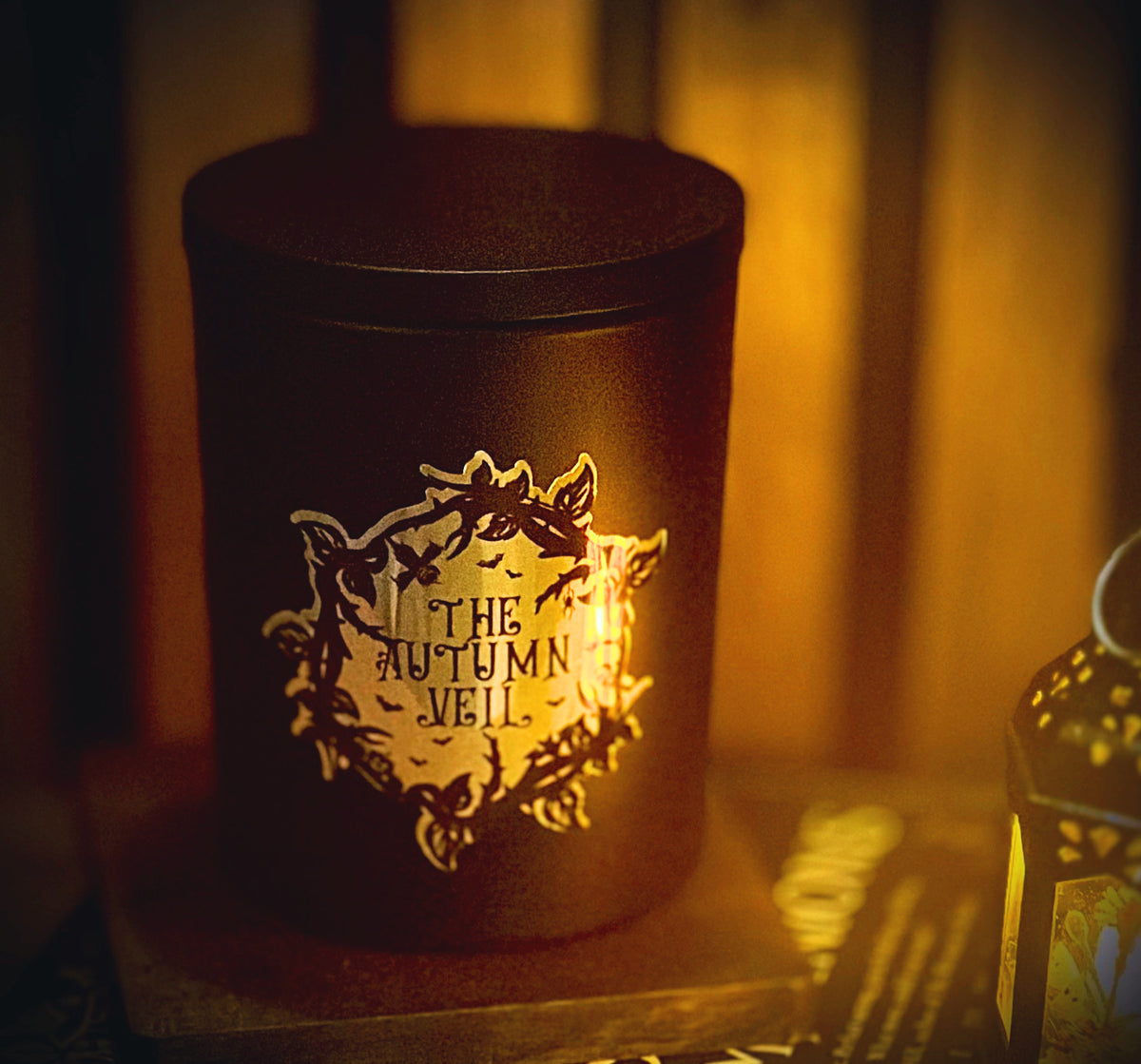 A dark cylindrical candle tin with a gold-toned decorative label reading 'THE AUTUMN VEIL,' photographed in warm amber lighting against a blurred indoor background.