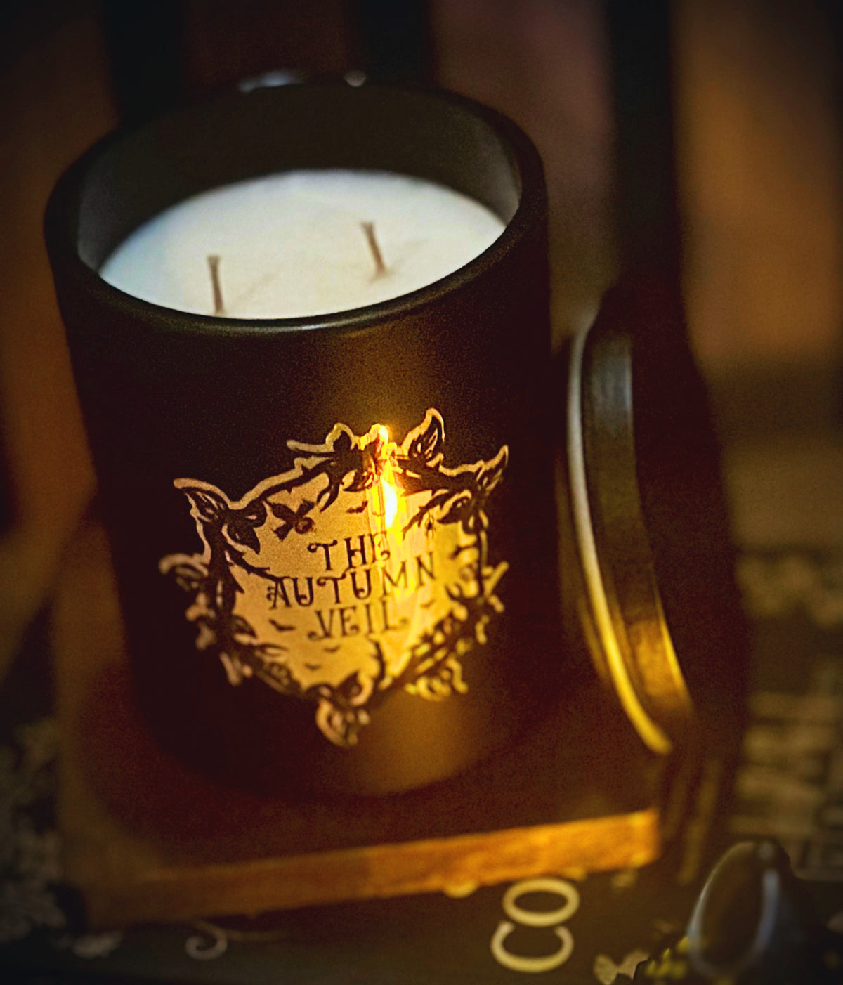 A black candle jar with a white wax fill and two wicks, featuring a gold foil crest and the text 'THE AUTUMN VEIL,' shown on a wooden surface with its lid beside it in a dimly lit setting.