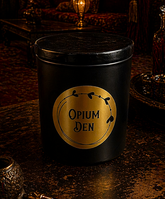 Matte black glass jar with black wooden lid and gold label, sitting on a dark wooden table beside a skull and dried roses. Soft candle glow emerging from under the lid.
