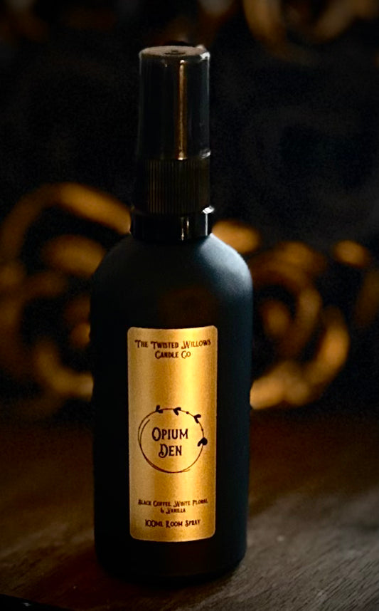 Opium Den room spray in a matte black glass bottle on a dark velvet cloth, next to scattered coffee beans and a dried floral bouquet.