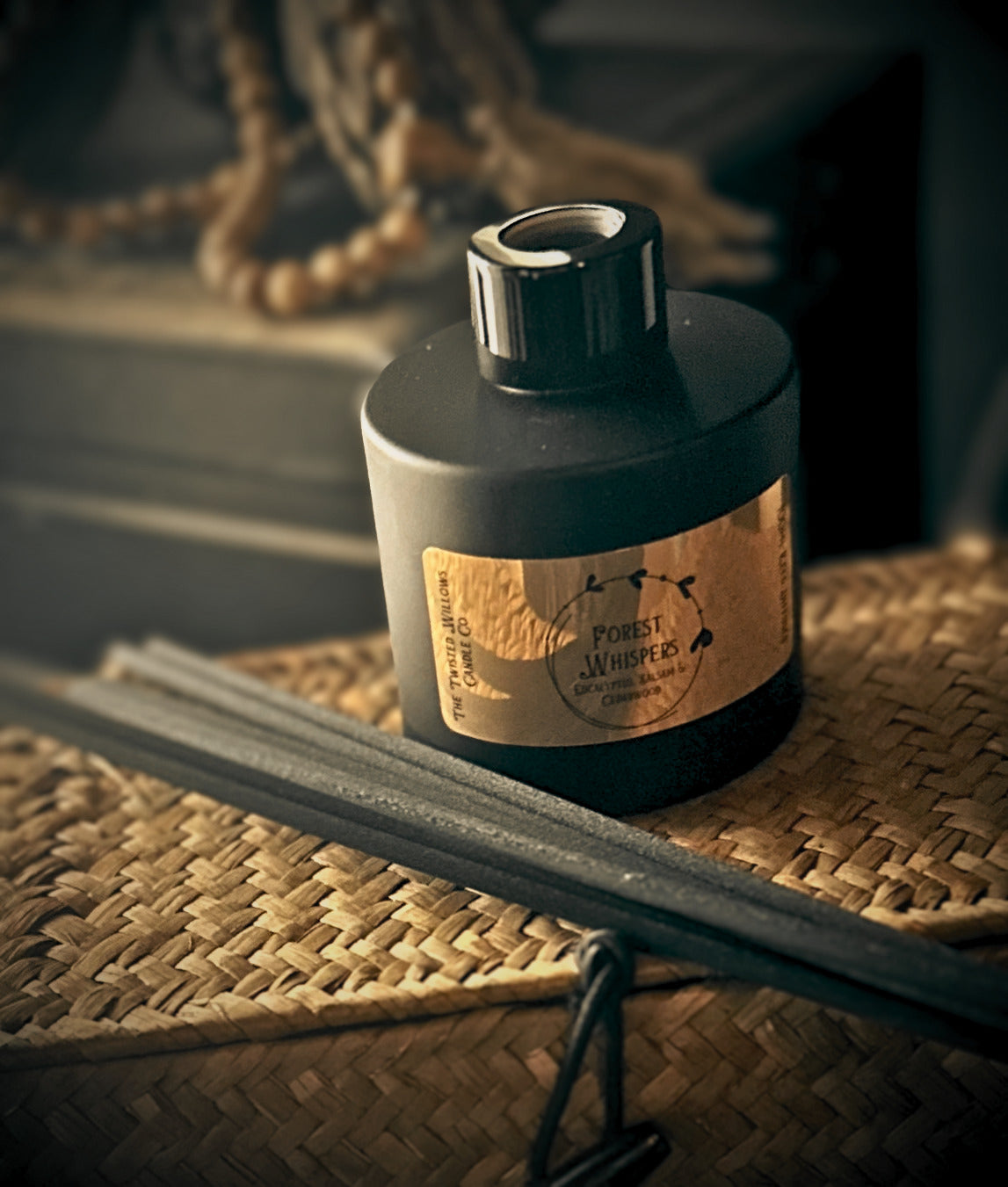 A black reed diffuser bottle with a gold label reading 'Forest Whispers' sits on a woven straw mat, with seven black reeds placed in front and a softly blurred rustic background.