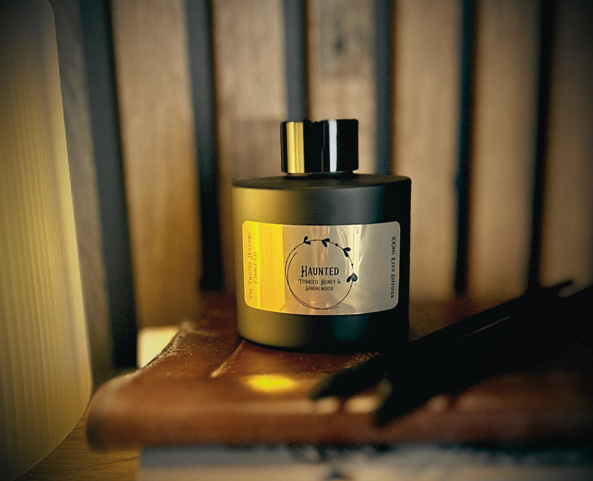A black reed diffuser bottle with a black cap and a gold  label reading 'HAUNTED'  placed on a wooden surface with a warm, dimly lit rustic background.