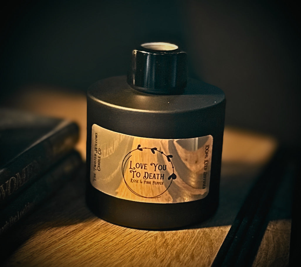A matte black diffuser bottle bottle with a black cap and a gold label reading 'LOVE YOU TO DEATH' , photographed on a wooden surface in warm, low-key lighting.